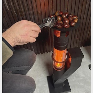 Könnte beinhalten: Ein schwarzer Metall-Holzofen mit einem transparenten Glaszylinder, der eine leuchtend orange Flamme zeigt. Ein runder Grill hält geröstete Kastanien. Zangen werden verwendet, um die Kastanien zu handhaben. Der Ofen steht auf einer Betonoberfläche.