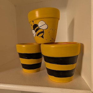 May include: Three yellow painted flower pots. Two pots have black stripes, and one pot has a bee design with a heart. The pots are on a white shelf. The pots are made of ceramic.