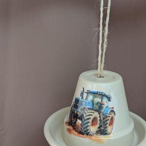 Peut inclure: Un pot de fleur suspendu, de couleur crème, orné d'un tracteur bleu. Le pot repose dans une soucoupe assortie et est suspendu par de la ficelle naturelle. Le tracteur a de gros pneus et se détache sur un fond marron.