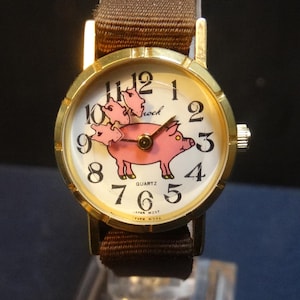 May include: A gold-toned wristwatch with a brown fabric band. The watch face features a pink pig illustration with the word "school" above it. The watch has black numerals and hands. The watch is displayed on a clear stand.