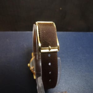 May include: A gold-toned wristwatch with a brown fabric band. The watch features a rectangular gold buckle and a clear watch face. The band has several holes for adjustment. The watch is a classic style.