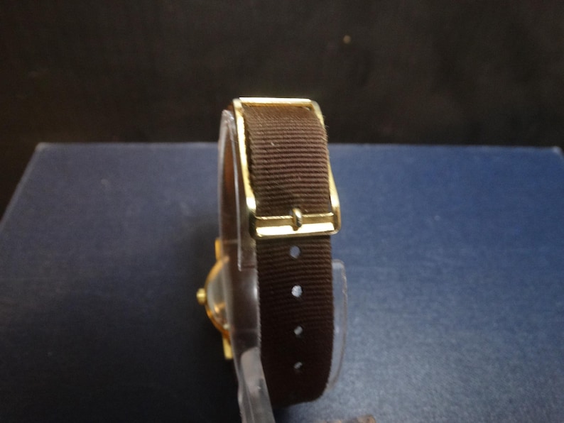 May include: A gold-toned wristwatch with a brown fabric band. The watch features a rectangular gold buckle and a clear watch face. The band has several holes for adjustment. The watch is a classic style.