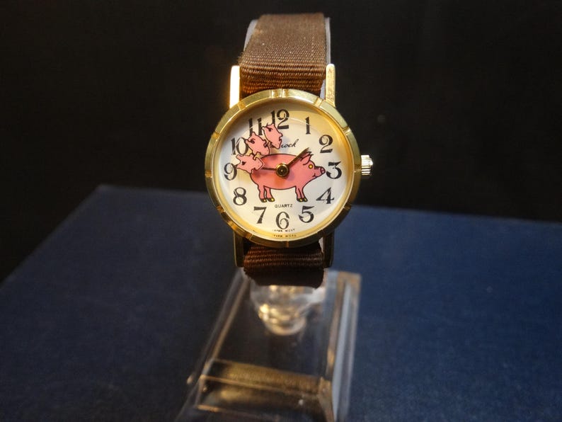 May include: A gold-toned wristwatch with a brown fabric band. The watch face features a pink pig illustration with the word "school" above it. The watch has black numerals and hands. The watch is displayed on a clear stand.