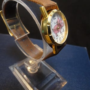 May include: A gold-toned wristwatch with a brown fabric band. The watch face features a pink pig illustration and white numerals. The watch is displayed on a clear stand, showcasing its design.
