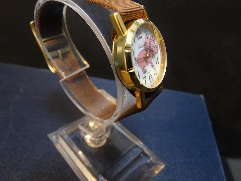 May include: A gold-toned wristwatch with a brown fabric band. The watch face features a pink pig illustration and white numerals. The watch is displayed on a clear stand, showcasing its design.