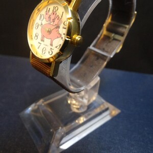 May include: A gold-toned wristwatch with a brown fabric band and a clear plastic strap. The watch face features a pink pig illustration with the numbers 1 through 12. The watch is displayed on a clear plastic stand.