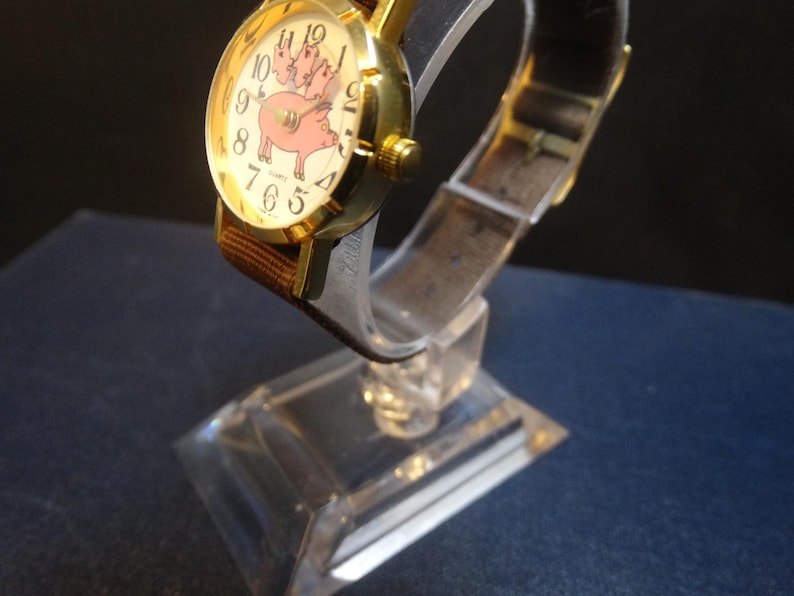 May include: A gold-toned wristwatch with a brown fabric band and a clear plastic strap. The watch face features a pink pig illustration with the numbers 1 through 12. The watch is displayed on a clear plastic stand.