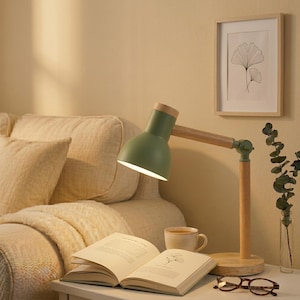May include: A green desk lamp with a wooden arm and base illuminates an open book. A framed botanical print and a cup of coffee sit nearby. The lamp's head is olive green, and the wooden arm extends to the base.