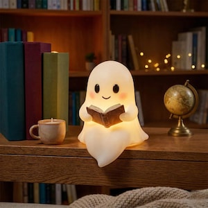 May include: A soft white ghost-shaped night light, holding a brown book, with a smiling face and rosy cheeks. A small candle in a mug and a miniature globe are on a wooden surface, with books in the background.