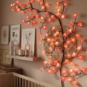 May include: A wall-mounted decorative cherry blossom branch with illuminated pink flowers. The branches are dark brown, and the flowers are lit with warm white lights. A woven circular wall hanging is behind the branch.