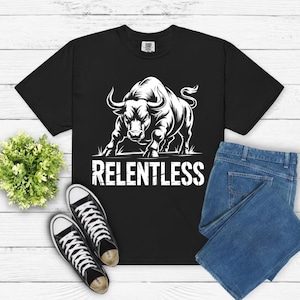 Peut inclure: T-shirt noir avec un graphique blanc d'un taureau et le mot "RELENTLESS". L'image comprend un jean bleu et des baskets noires et blanches. Une petite plante verte est sur le côté gauche de l'image.