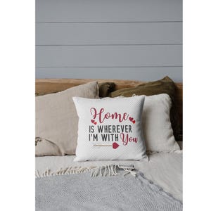 Puede incluir: Un cojín cuadrado blanco con el texto "Home is wherever I'm with You" en escritura roja y un diseño de corazón rojo. El cojín está sobre una cama con otras almohadas y una manta gris.