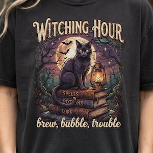Peut inclure: Un t-shirt noir avec un motif représentant un chat noir assis sur une pile de livres intitulés Spells, Potions et Lore. À côté du chat se trouve une lanterne allumée. Le texte indique Witching Hour en haut et brew, bubble, trouble en bas.