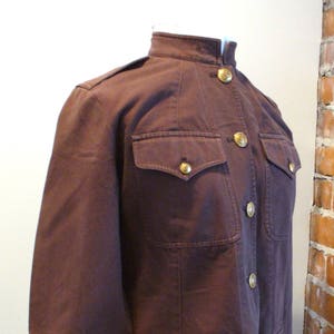 May include: A dark brown jacket with a mandarin collar, four front pockets, and gold-colored buttons. The jacket has epaulets on the shoulders and a tailored fit. The garment is displayed on a mannequin.