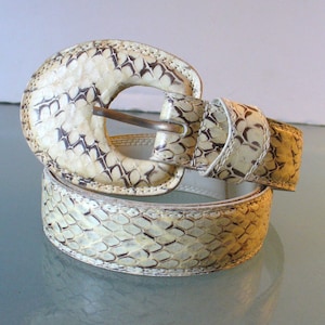 Könnte beinhalten: Ein cremefarbener Gürtel mit Schlangenmuster. Der Gürtel hat eine große, ovale Schnalle mit passendem Schlangenhaut-Design und einen messingfarbenen Metallverschluss. Der Gürtel ist auf einer reflektierenden Oberfläche aufgerollt.