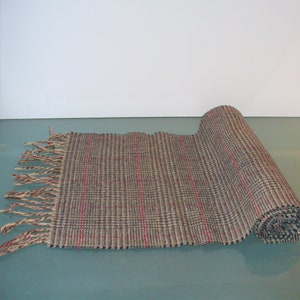 May include: A rectangular woven scarf featuring a plaid pattern in shades of brown, blue, and grey with thin vertical red stripes. The scarf is partially rolled at one end and finished with long, loose fringe tassels along the opposite edge.