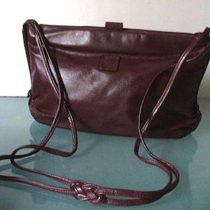 May include: A dark burgundy leather shoulder bag featuring a rectangular silhouette, a smooth finish, and a thin adjustable strap with a decorative knot detail. The bag includes a central flap closure with a small rectangular tab and a square patch.