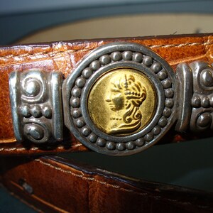 Pu&ograve; includere: Primo piano di una cintura in pelle marrone con fibbia argentata e dorata. La fibbia presenta un centro dorato a forma di moneta con il profilo di una persona, circondato da perline argentate e dettagli decorativi argentati.