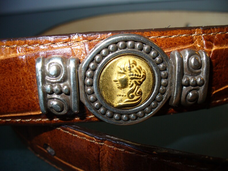Pu&ograve; includere: Primo piano di una cintura in pelle marrone con fibbia argentata e dorata. La fibbia presenta un centro dorato a forma di moneta con il profilo di una persona, circondato da perline argentate e dettagli decorativi argentati.