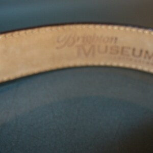 May include: Close-up of a brown leather belt with the words "Brighton MUSEUM" embossed on the inside. The belt has a stitched edge and a light tan interior. The background is a muted teal color.