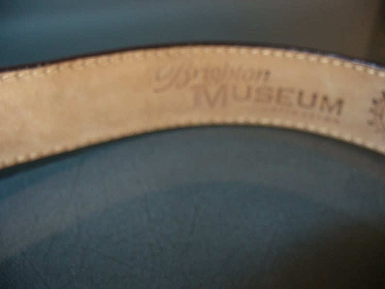 May include: Close-up of a brown leather belt with the words "Brighton MUSEUM" embossed on the inside. The belt has a stitched edge and a light tan interior. The background is a muted teal color.