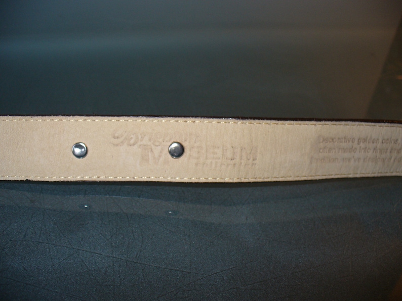 Pu&ograve; includere: Primo piano di una cintura in pelle marrone con rivetti color argento. L'interno della cintura &egrave; di colore beige chiaro con testo in rilievo. La cintura fa parte della "Museum Collection".