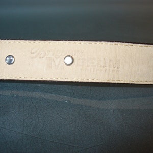 Pu&ograve; includere: Primo piano dell'interno di una cintura in pelle marrone, con due rivetti argentati e testo in rilievo. La fodera color crema mostra il nome del marchio e la taglia. Le cuciture sono visibili sui bordi.