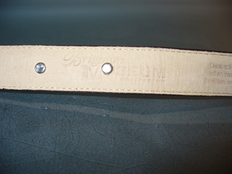 Pu&ograve; includere: Primo piano dell'interno di una cintura in pelle marrone, con due rivetti argentati e testo in rilievo. La fodera color crema mostra il nome del marchio e la taglia. Le cuciture sono visibili sui bordi.