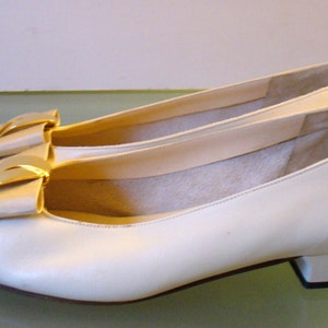 Può includere: Un paio di ballerine in pelle color crema con un piccolo tacco. Ogni scarpa presenta un fiocco decorativo con un accento dorato. Le scarpe sono progettate per un look classico ed elegante.