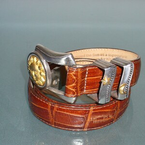 May include: Brown leather belt with a textured crocodile pattern. The belt buckle is silver-toned with a gold sun design. Additional silver-toned belt loops feature a star and crescent moon.