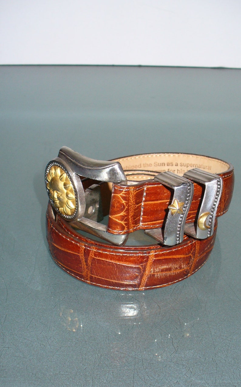 May include: Brown leather belt with a textured crocodile pattern. The belt buckle is silver-toned with a gold sun design. Additional silver-toned belt loops feature a star and crescent moon.