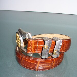 May include: A brown leather belt with a textured, crocodile-like pattern. The belt features a silver buckle with a gold flower design and two additional silver belt loops, one with a star and one with a crescent moon.