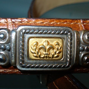 Pu&ograve; includere: Primo piano di una cintura in pelle marrone con una fibbia decorativa argentata e dorata. La fibbia presenta un disegno a giglio dorato all'interno di una cornice argentata. La cintura ha una superficie strutturata.