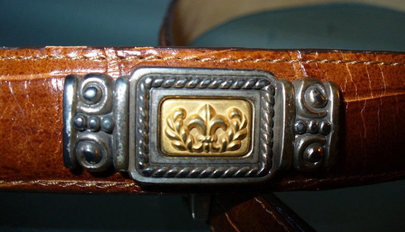 Pu&ograve; includere: Primo piano di una cintura in pelle marrone con una fibbia decorativa argentata e dorata. La fibbia presenta un disegno a giglio dorato all'interno di una cornice argentata. La cintura ha una superficie strutturata.