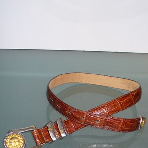 May include: A brown leather belt with a textured, crocodile-like pattern. The belt features a silver buckle with a gold sun design and silver accents. The brand name "Brighton MUSE" is visible on the inside of the belt.