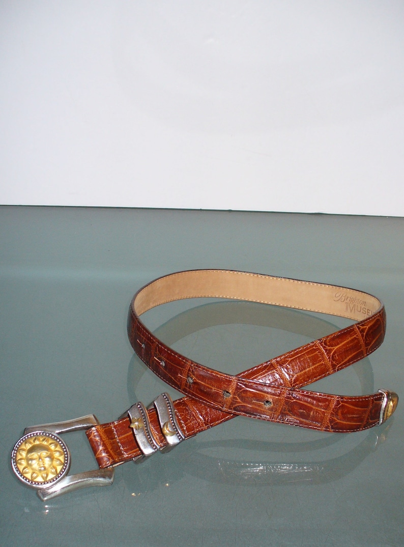 May include: A brown leather belt with a textured, crocodile-like pattern. The belt features a silver buckle with a gold sun design and silver accents. The brand name "Brighton MUSE" is visible on the inside of the belt.