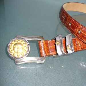 May include: A brown leather belt with a silver buckle featuring a gold sun design. The belt has silver accents with a star and crescent moon. The belt is approximately 1.5 inches wide.