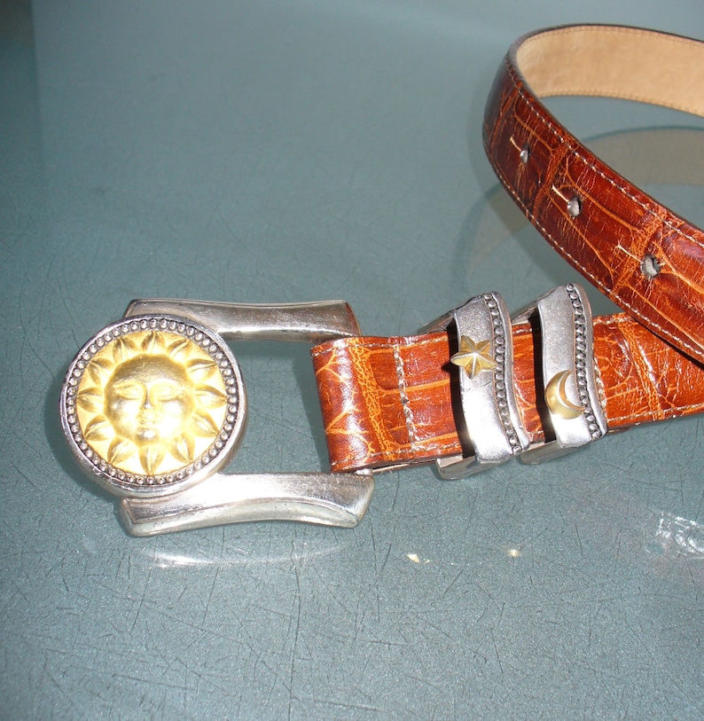 May include: A brown leather belt with a silver buckle featuring a gold sun design. The belt has silver accents with a star and crescent moon. The belt is approximately 1.5 inches wide.