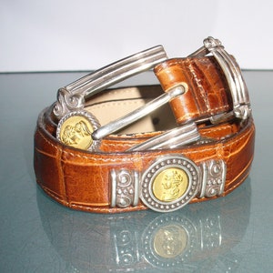 Pu&ograve; includere: Una cintura in pelle marrone con texture a coccodrillo. La cintura presenta elementi metallici argentati e medaglioni dorati. La fibbia e i passanti sono anch'essi argentati. La cintura &egrave; arrotolata su una superficie riflettente.