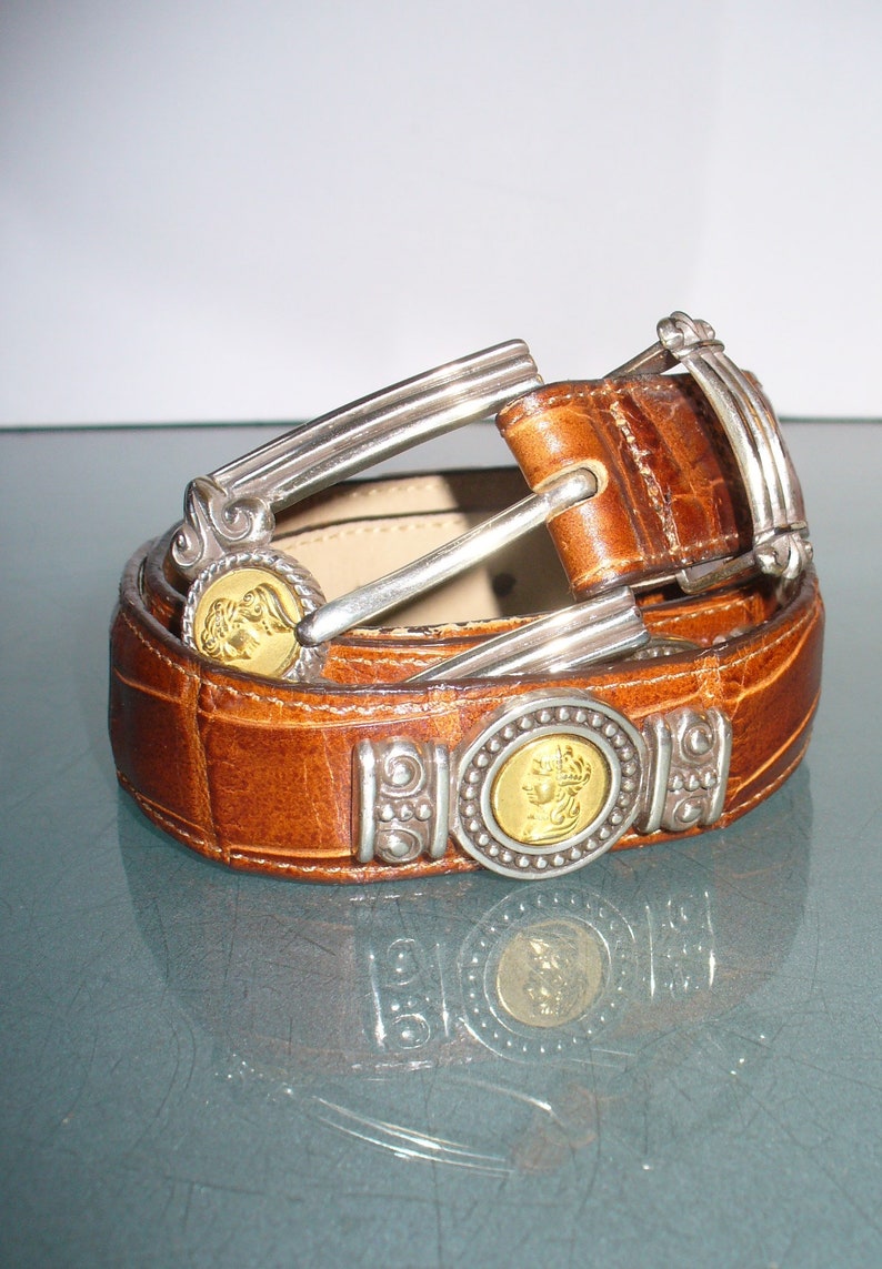 Pu&ograve; includere: Una cintura in pelle marrone con texture a coccodrillo. La cintura presenta elementi metallici argentati e medaglioni dorati. La fibbia e i passanti sono anch'essi argentati. La cintura &egrave; arrotolata su una superficie riflettente.