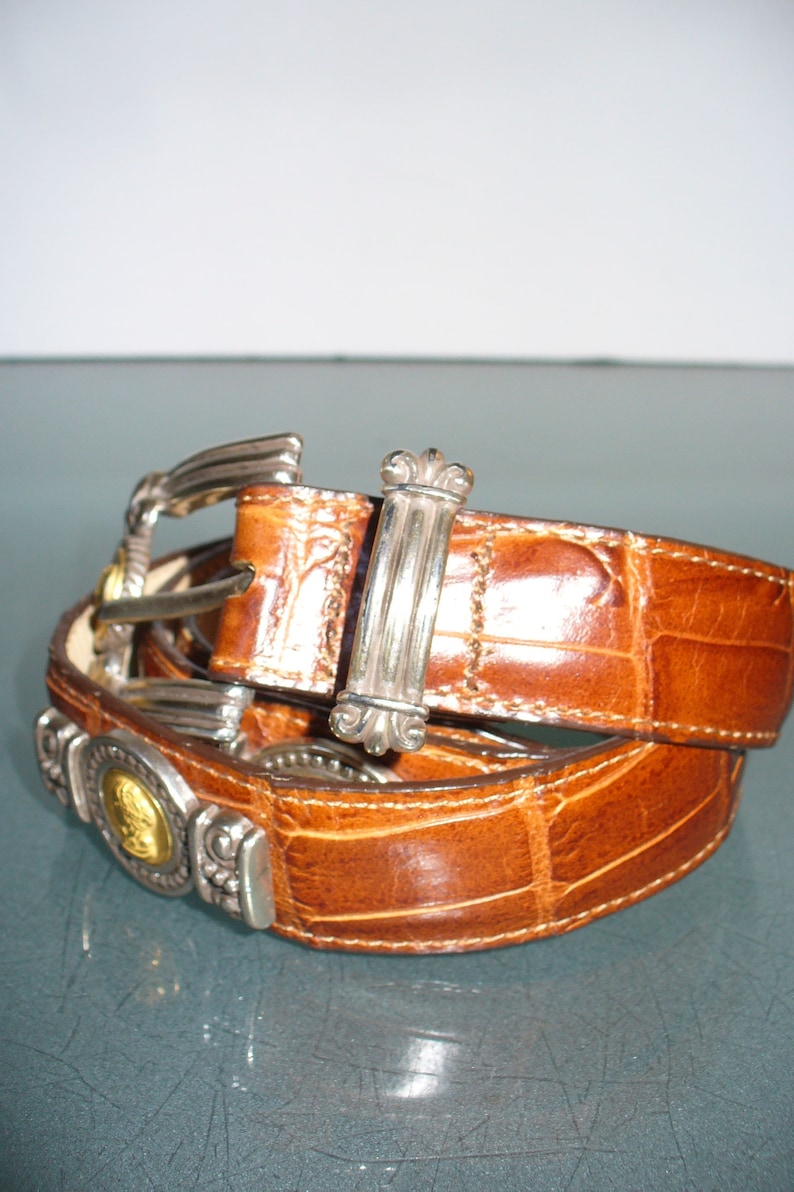 Pu&ograve; includere: Una cintura in pelle marrone con motivo a pelle di coccodrillo. La cintura presenta una fibbia argentata e dettagli decorativi argentati e dorati. La cintura &egrave; ripiegata su se stessa.