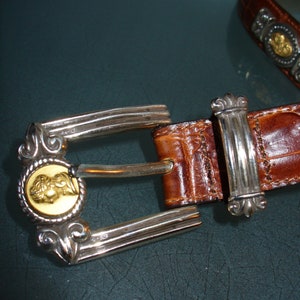 Pu&ograve; includere: Una cintura in pelle marrone con fibbia argentata. La fibbia presenta un medaglione dorato con un design dettagliato. La cintura ha un motivo strutturato a forma di coccodrillo.