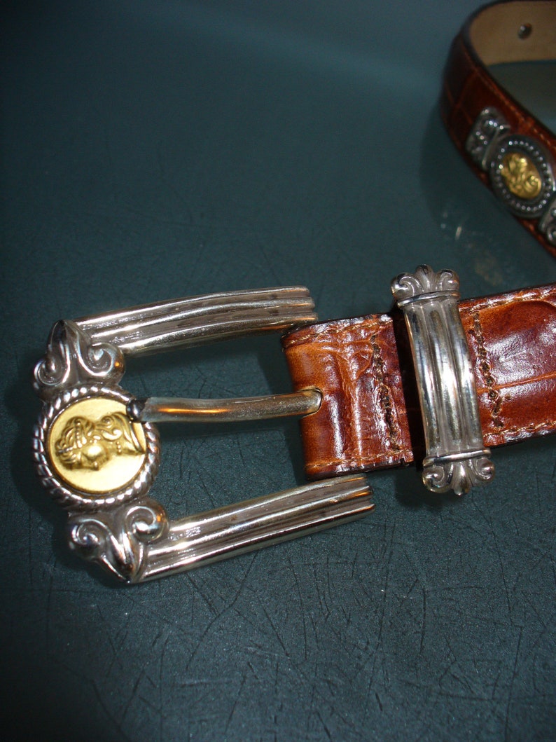 Pu&ograve; includere: Una cintura in pelle marrone con fibbia argentata. La fibbia presenta un medaglione dorato con un design dettagliato. La cintura ha un motivo strutturato a forma di coccodrillo.