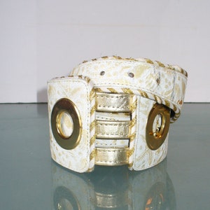 May include: White belt with gold accents. The belt features a repeating floral pattern and gold metal rings. The belt has a gold metallic trim and gold metallic accents. The belt is a fashion accessory.