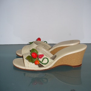 May include: A pair of cream-colored wedge sandals with woven raffia straps. The sandals are decorated with red and orange fruit and green leaves. The wedge heels are made of cork.
