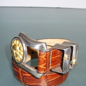 May include: Brown leather belt with a textured pattern. The belt features a silver buckle with a gold sun face design and two additional silver and gold accents. The belt is a fashion accessory.