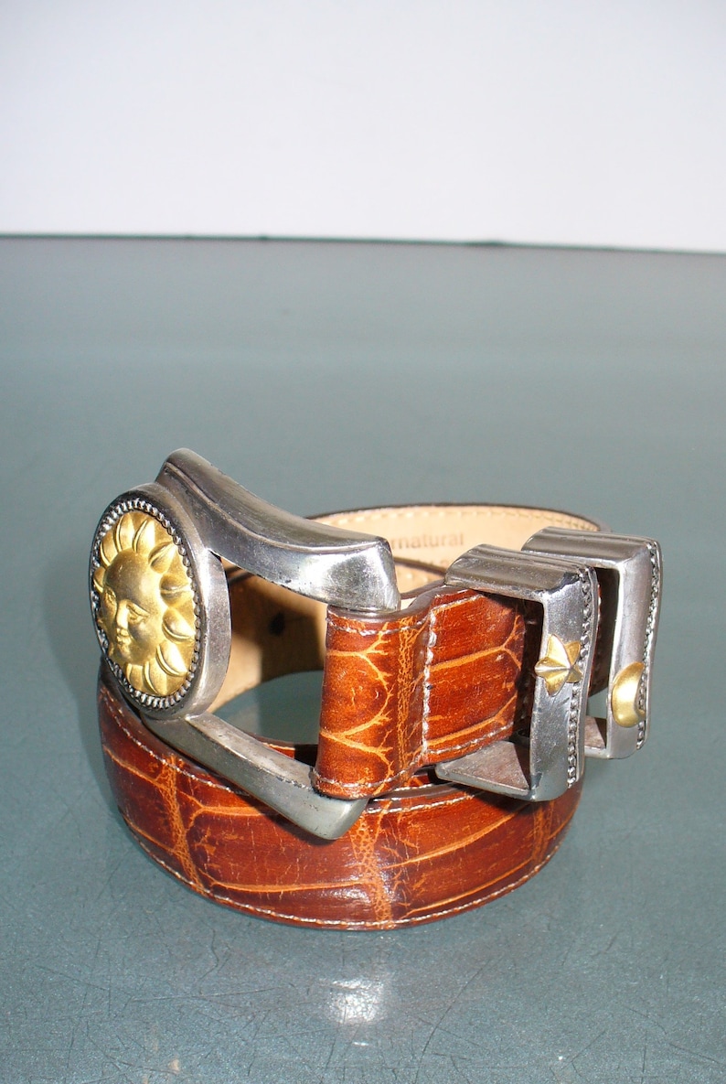 May include: Brown leather belt with a textured pattern. The belt features a silver buckle with a gold sun face design and two additional silver and gold accents. The belt is a fashion accessory.