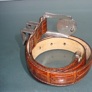 May include: A brown leather belt with a textured, crocodile-like pattern. The belt features a silver-colored buckle with a unique design. The belt is coiled, showcasing its full length and the details of the leather.