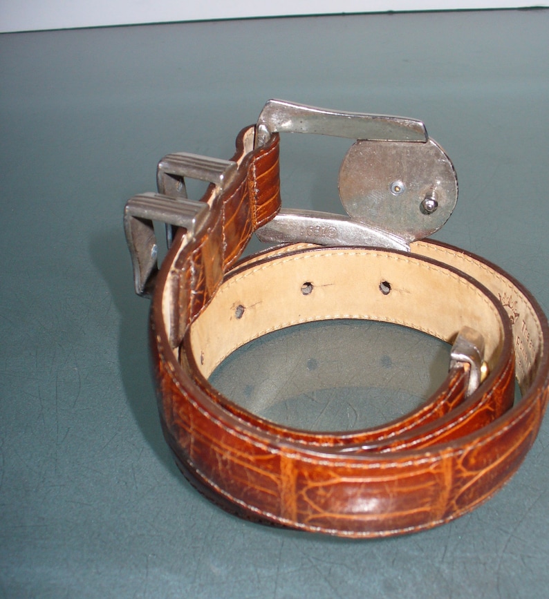 May include: A brown leather belt with a textured, crocodile-like pattern. The belt features a silver-colored buckle with a unique design. The belt is coiled, showcasing its full length and the details of the leather.