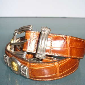 Pu&ograve; includere: Cintura in pelle marrone con motivo coccodrillo. La cintura presenta dettagli in metallo argentato, tra cui una fibbia, placche decorative e un passante rettangolare. &Egrave; presente anche un medaglione dorato.